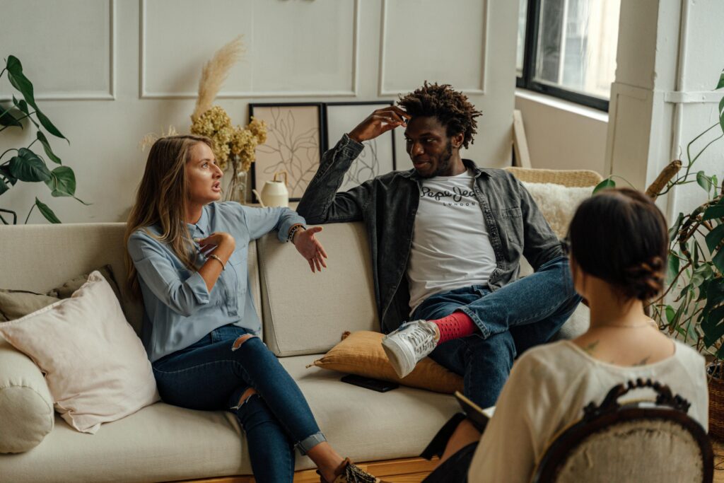 Three people are engaged in conversation, seated on a sofa in a well-lit room with plants and wall art.